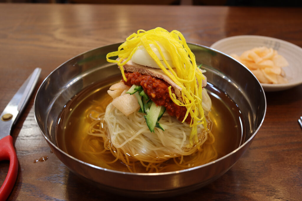 밀면 (Busan Milmyeon / Busan cold wheat noodles) — a representative photo of the dish, not necessarily from the restaurants above. Photo: Photo by Mobius6, via Wikimedia Commons, licensed CC BY-SA 4.0 (https://creativecommons.org/licenses/by-sa/4.0)