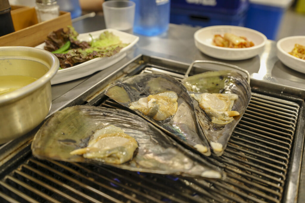 조개구이 (Jogae-gui / grilled shellfish) — a representative photo of the dish, not necessarily from the restaurants above. Photo: Photo by 'bryan...' (Flickr: bryansjs), via Wikimedia Commons. Licensed under CC BY-SA 2.0 (https://creativecommons.org/licenses/by-sa/2.0). Source: https://commons.wikimedia.org/wiki/File:Jogae-gui_3.jpg (original https://www.flickr.com/photos/bryansjs/43931345240/)