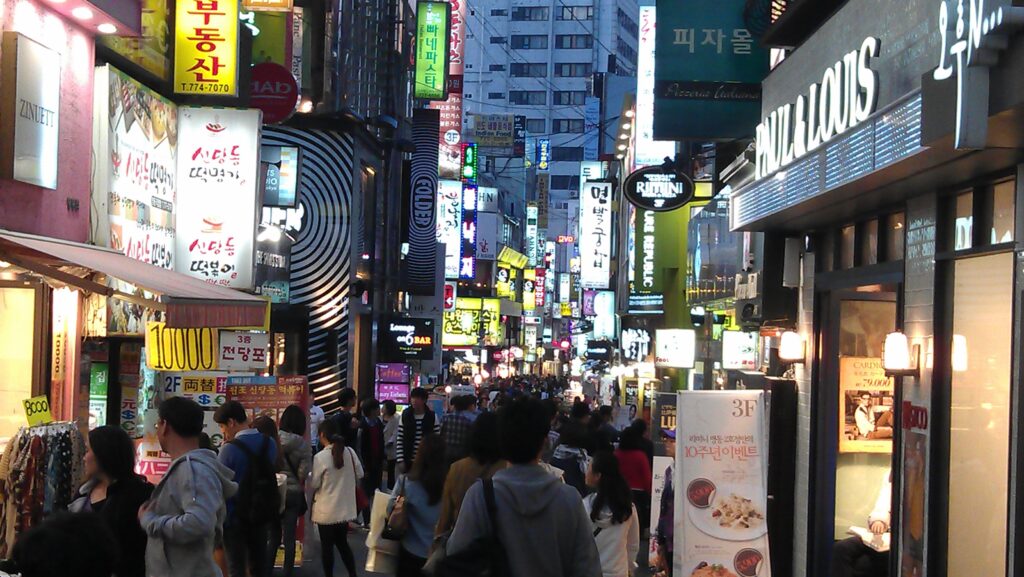 Neon-lit night streets of Seoul's Myeongdong district — illustrative of the moody neon-noir Seoul cityscape evoked by the series, not a confirmed filming location. (Photo: Christian Bolz, CC BY-SA 4.0 via Wikimedia Commons)