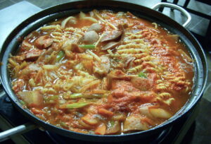 A steaming pot of Korean budae-jjigae army base stew with sausage, Spam, kimchi and noodles in a spicy red broth