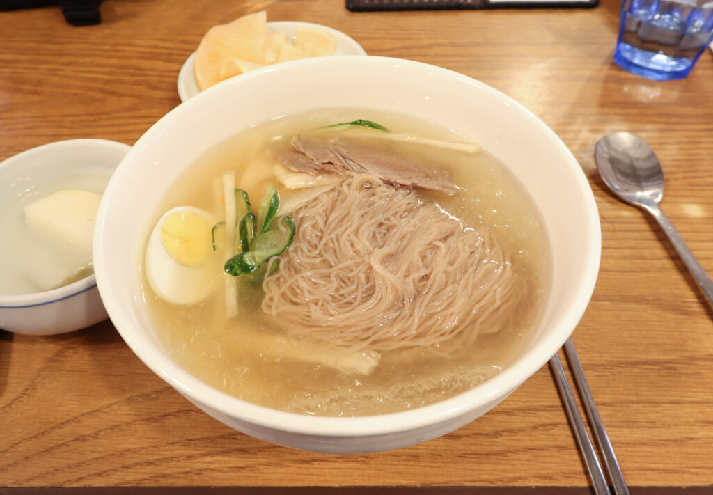 Pyongyang-style naengmyeon, with high-buckwheat noodles in a clear, restrained beef broth meant to be tasted plain first. (Photo: Mobius6, CC BY-SA 4.0, via Wikimedia Commons)