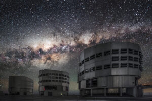 The Milky Way stretching across a star-filled night sky above silhouetted telescope domes of an observatory